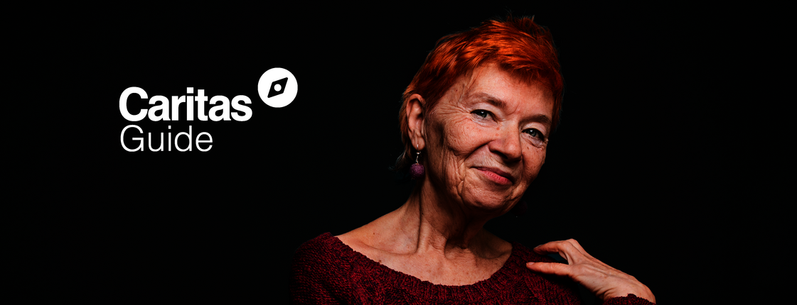 Elderly woman with short hair smiling into the camera, the background is black. Logo Caritas Guide in the top left corner. 